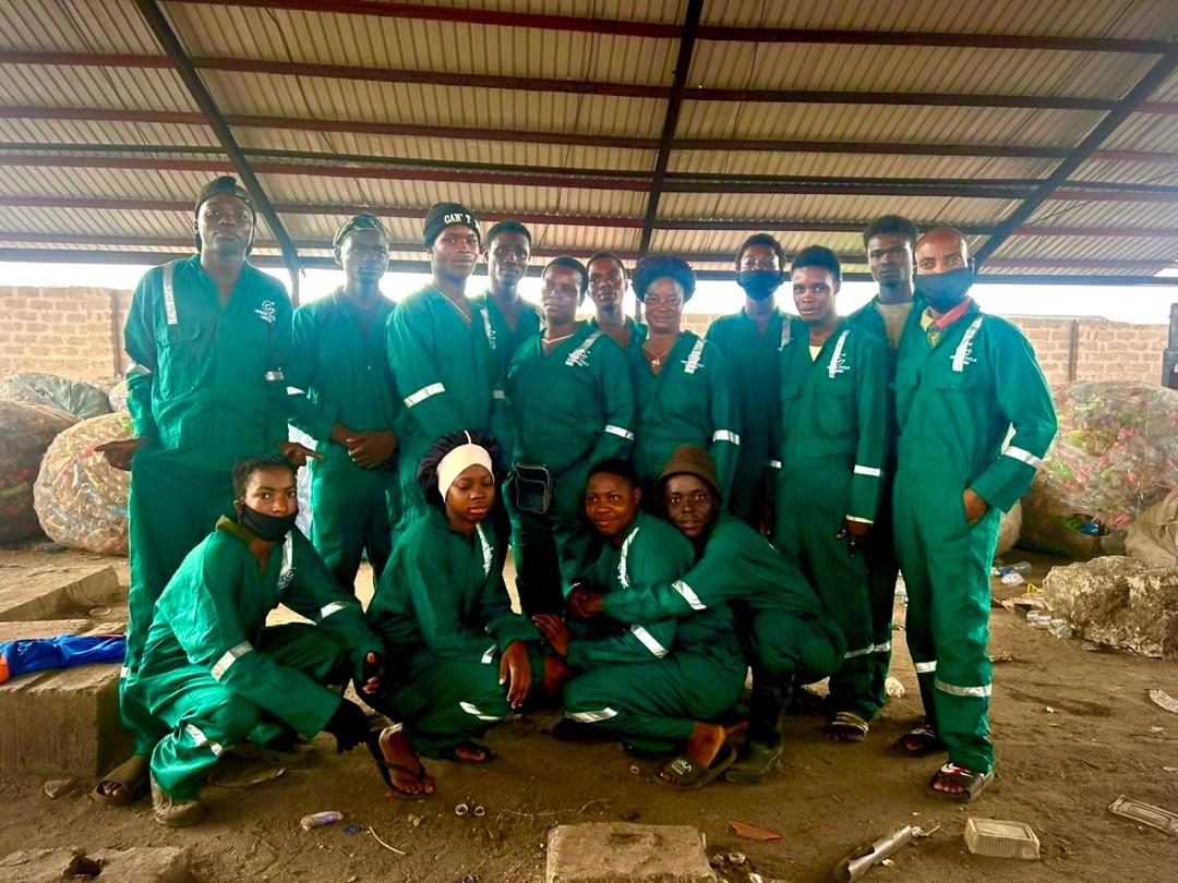 The Green Cycler operations team in green overalls with reflective strips, standing and kneeling in the factory shed with baled PET bottles behind them